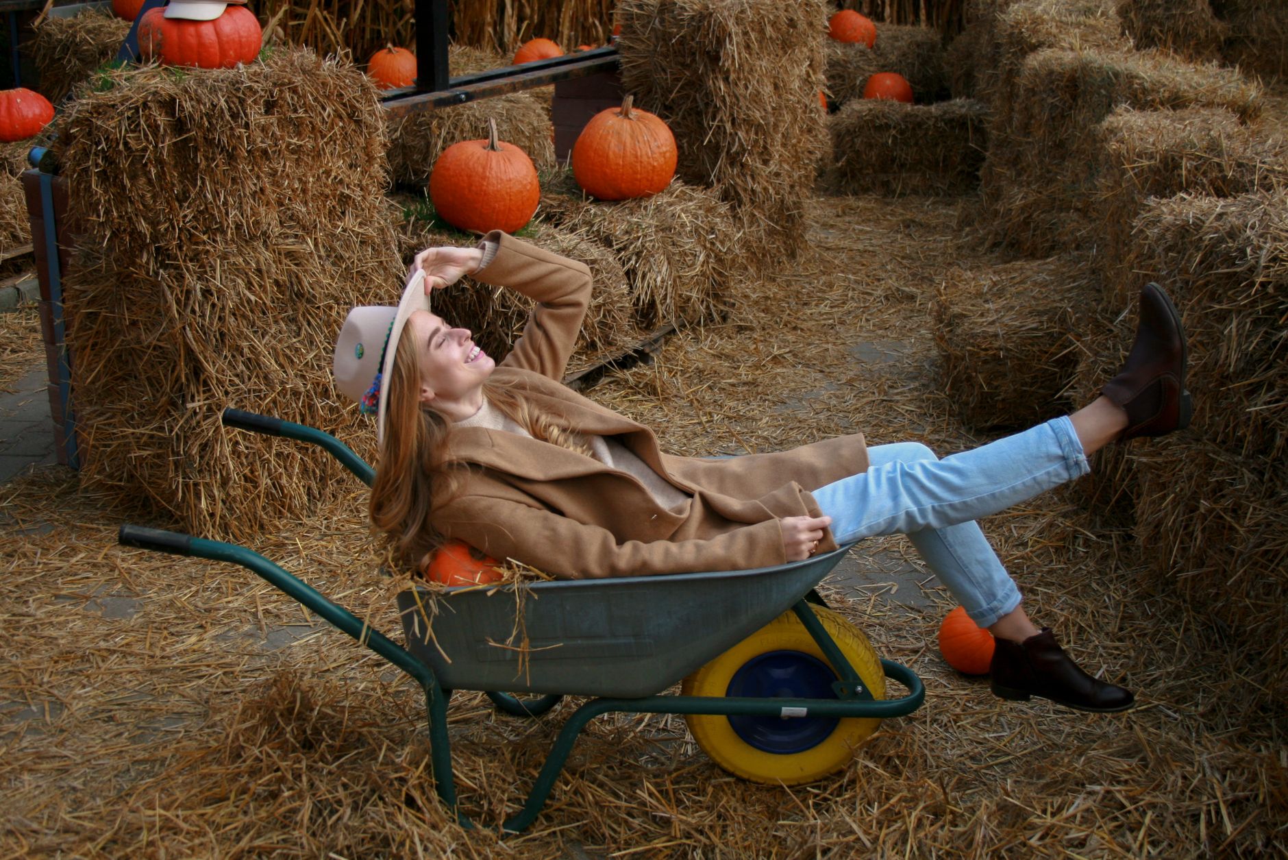 A woman in a coat and hat reclines in a wheelbarrow surrounded by hay and pumpkins on a farm.
