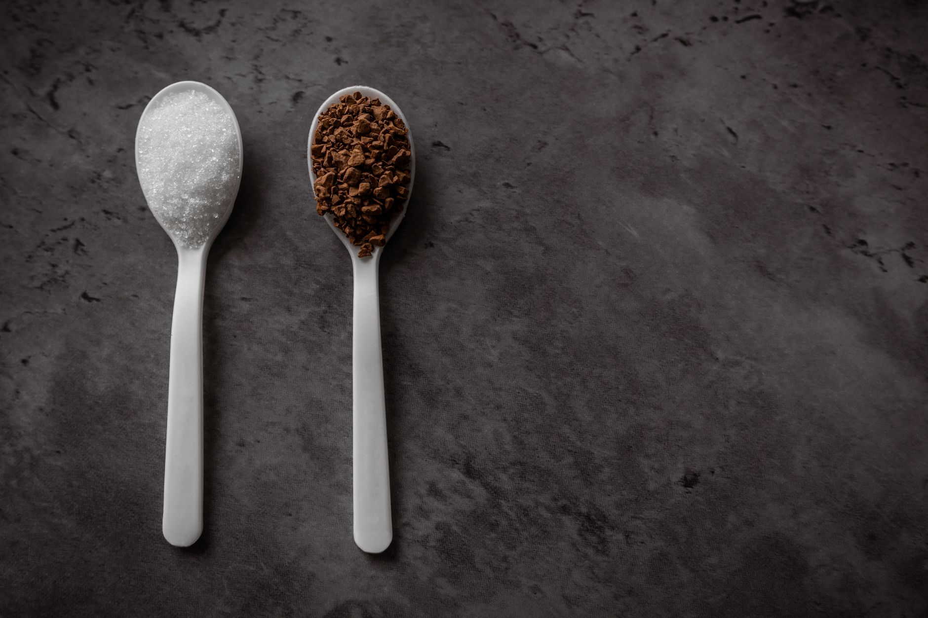Close-up of sugar and coffee granules on spoons against a dark textured surface.