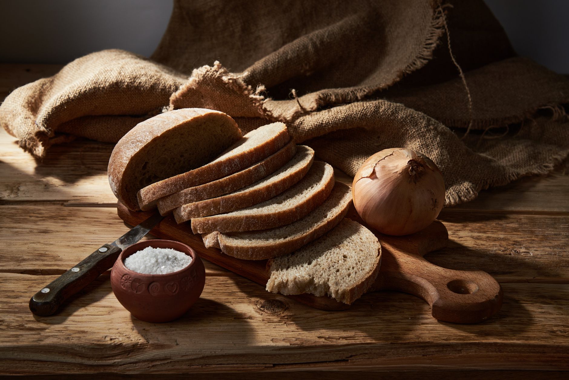 A still life composition featuring sliced artisan bread accompanied by an onion and a bowl of salt on a rustic wooden board.